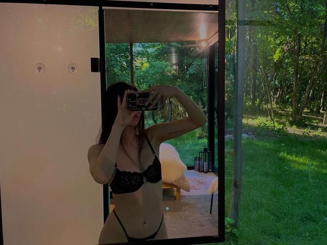 CherryMuse – in einem schwarzen Spitzen-BH und String, hält ein Handy, Selfiemit Spiegel, zeigt ihre Brüste und Vulva, steht vor einer Glastür, draußen ein Wald, Sonnenlicht..