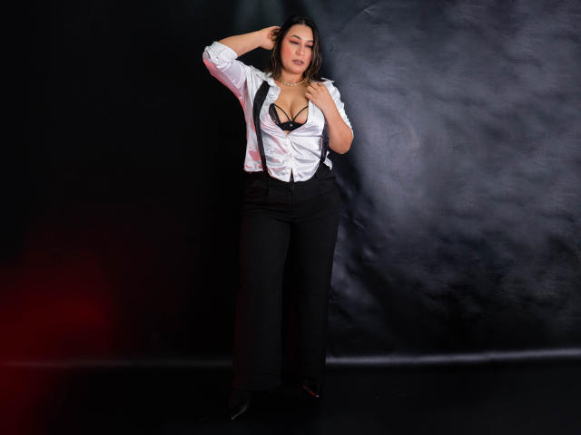 EmaFoxy, con un seno di media grandezza, indossa una camicia bianca, pantaloni neri e tacchi neri, è in piedi davanti a uno sfondo scuro, con una mano tra i capelli, e indossa un reggiseno nero sotto i vestiti..