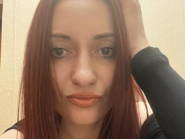 Izzy Kelley, com cabelo ruivo, mão dentro de uma manga preta, usando batom laranja, olhos castanhos e um leve sorriso, olhando diretamente para a câmera. Ela tem um tom de pele claro..