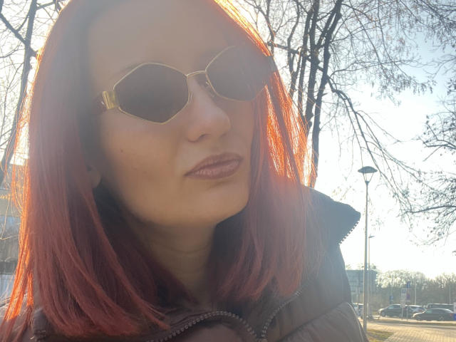 Izzy Kelley met rood haar, die een aviatorzonnebril draagt en een dikke zwarte jas, met zonlicht op de achtergrond, die licht glimlacht met glanzende bruine lippen, in een buitenomgeving..