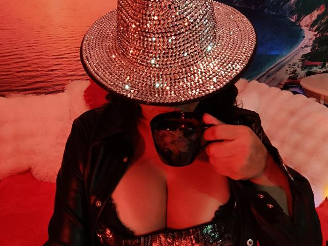 AmberSunshine with big tits in a black sequined top, holding a black lace mask, wearing a sparkly silver hat, red lighting, black leather jacket, and sipping from a cup.