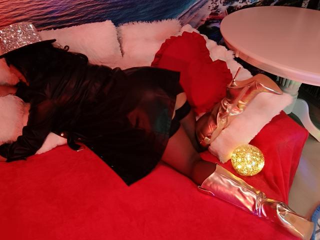 AmberSunshine ligt op een rode bed, gekleed in een zwarte jurk, zwarte kousen en glanzende gouden laarzen, met een glitterbal in de buurt van haar achterkant, en een witte ronde tafel..