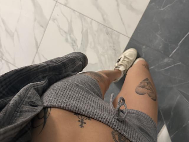 SensualMila's thighs, tattooed with a heart and an ankh, in gray ribbed shorts, white sneakers, and an open gray shirt, standing on a marble floor.