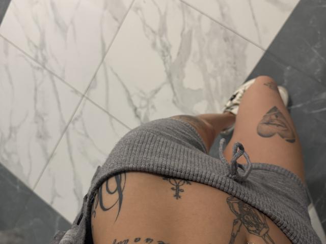 Les fesses de SensualMila, recouvertes d'une jupe gris à rayures, laissant apparaître des tatouages sur le bas du dos et la cuisse. Elle porte des baskets blanches et noires. Un sol en marbre se trouve en dessous..