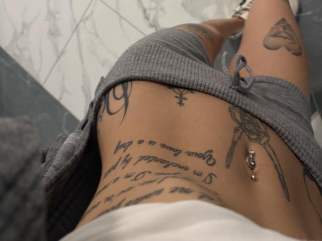 La parte inferiore dell'addome di SensualMila, i tatuaggi, i pantaloncini grigi a coste, il piercing all'ombelico, il tatuaggio a forma di cuore visibile sulla coscia, ripreso dall'alto. La pelle abbronzata di SensualMila, i tatuaggi e i pantaloncini dominano l'inquadratura..