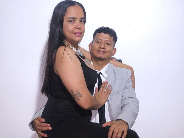 LilithLuciffer, eine Latina mit langen schwarzen Haaren, kleinen Brüsten, einem schwarzen Kleid und Tattoos, sitzt auf einem etwas korpulenten Latino in einem grauen Anzug, weißem Hemd und rotem Krawatte, der seine Brust festhält und lächelt..