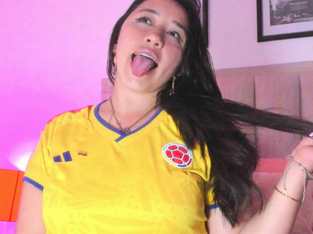 JessyBarnnett with long dark hair, tongue out, wearing a yellow soccer shirt with blue trim and a red soccer ball emblem, holding her hair. Pink light in background.