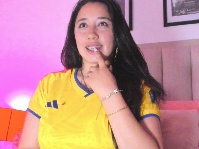 Jessy Barnnett mit langen, schwarzen Haaren, einem gelben Hemd mit blauer Verzierung, einem Finger im Mund, einem Lächeln, einem goldenen Armband, rosafarbenem und orangefarbenem Licht und einem beigen Kopfteil im Hintergrund..