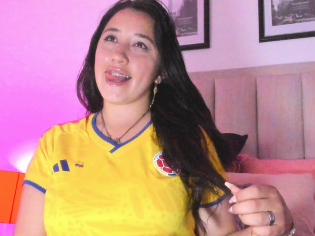 JessyBarnnett with long dark hair, yellow soccer shirt, blue trim, and red logo, sticking out tongue, holding cigarette, pink and purple lighting, smiling, wearing gold earrings.