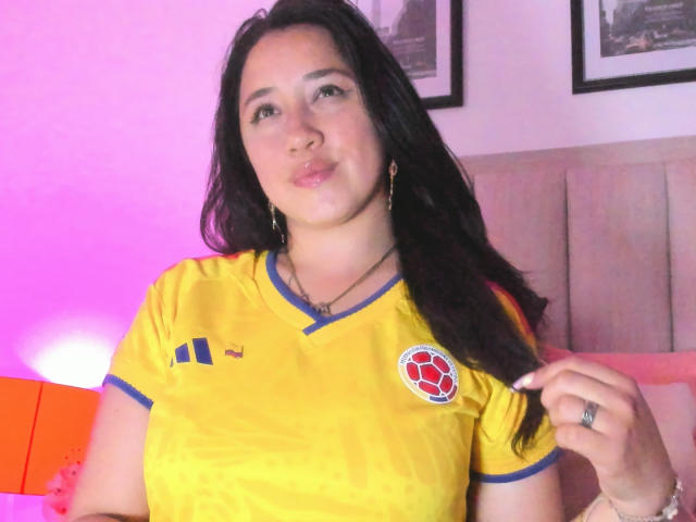 JessyBarnnett with long black hair, wearing a yellow soccer shirt with blue trim and a red soccer logo, pouting her lips, holding her hair, and wearing a ring.