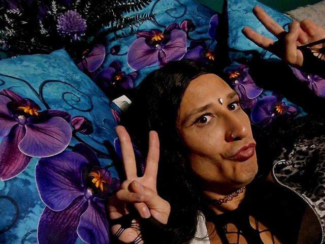 MaryXXL-hot with dark hair, nose ring, and choker, lying on blue floral pillow, making a double peace sign, wearing a black lace-up top, with a serious expression.