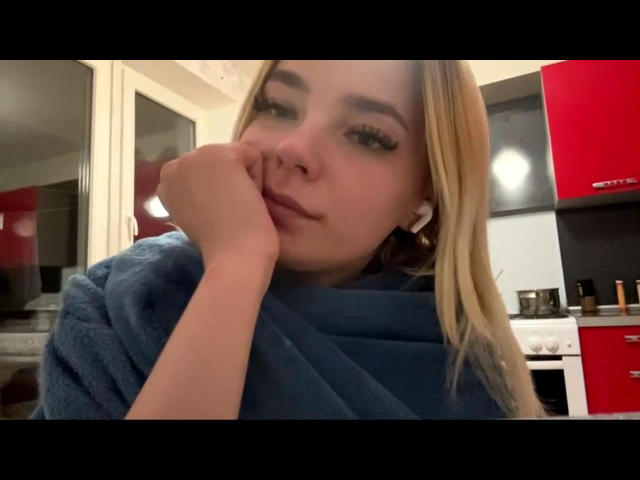 Adriene Degennaro, com cabelo loiro, usando um lenço azul, com o queixo apoiado na mão, olhando ligeiramente para o lado, em uma cozinha com armários vermelhos e pretos..