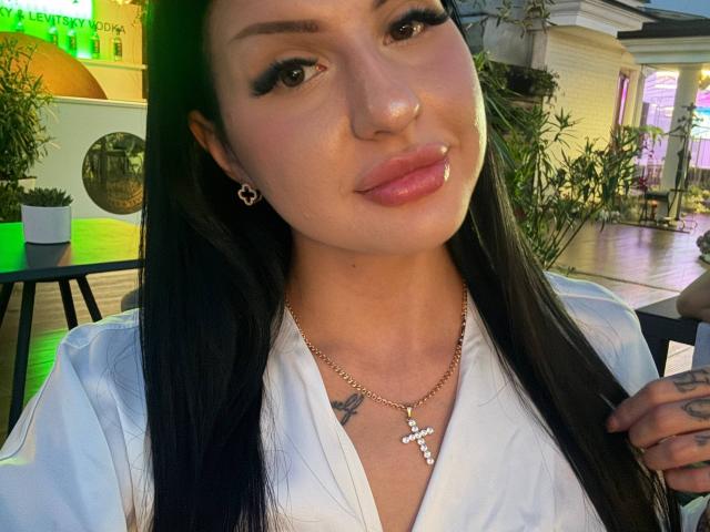 AuraXo, com cabelos longos e pretos, olhos castanhos, piercing no nariz e no lábio, vestindo uma camisa branca, um colar com uma cruz e brincos em formato de estrela, sorrindo para a câmera..