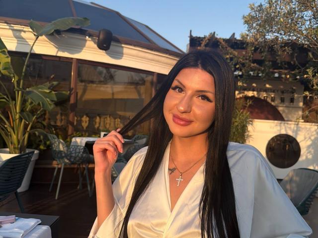 AuraXo, avec de longs cheveux noirs, tenant une clé, portant une robe blanche, un collier avec une croix, souriante, en plein soleil, sur un toit, avec des plantes, des chaises en métal, en fin d'après-midi, lumière chaude, séduisante..