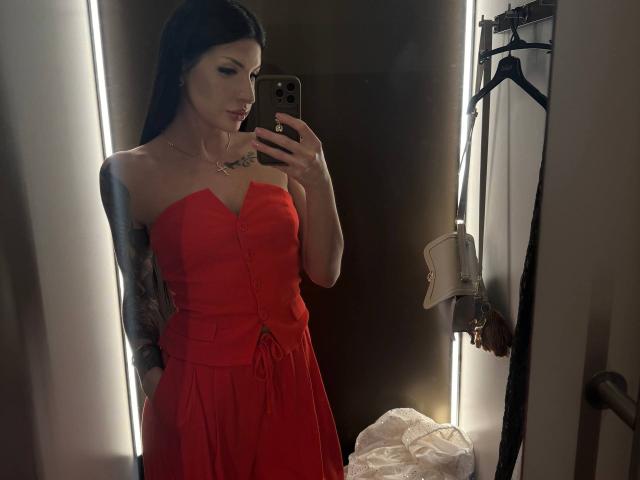 AuraXo, con cabello negro, vestida con un vestido rojo sin tirantes, sosteniendo un teléfono en un selfie frente a un espejo, mostrando tatuajes en su brazo y un collar con un diseño de estrella..