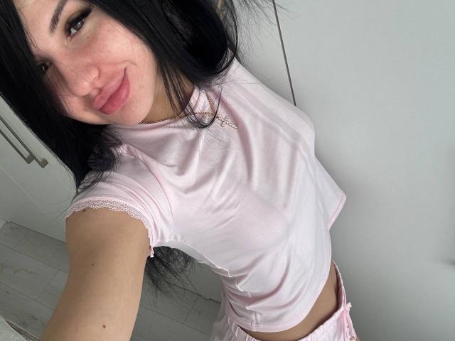 AuraXo, avec de longs cheveux noirs, un t-shirt rose à dentelle, de petites seins, un pantalon rose, souriante, peau claire, porte une porte blanche, parquet en bois, un collier discret, un selfie décontracté..
