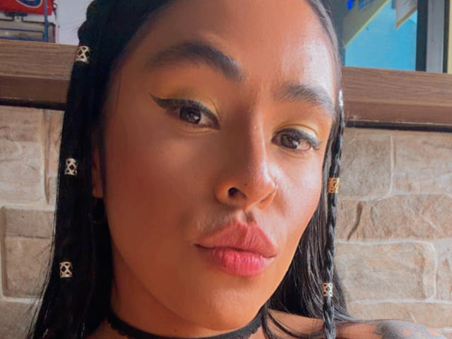 StellaBeckker's face with dark braids, yellow eyeshadow, winged eyeliner, black choker, and pouty pink lips. Her brown skin is shiny, and she's making a kissy face.
