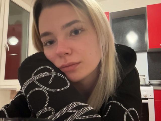 Adriene Degennaro, con i capelli biondi, indossa una felpa nera con motivi circolari bianchi, appoggiata a un bancone della cucina, con un'aria leggermente annoiata, con un sorriso appena accennato sul viso..