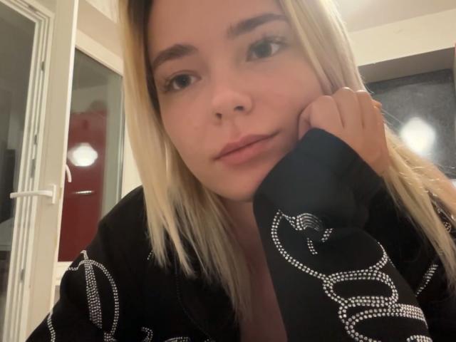 Adriene Degennaro, com cabelo loiro, olhos castanhos e pele clara, apoiando o queixo em uma manga preta com padrões brancos, olhando para a câmera com um leve sorriso..