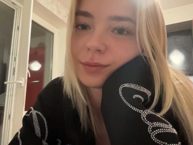 Adriene Degennaro, con i capelli biondi, indossa una veste nera con motivi a spirale bianchi, appoggia la testa sulla mano, sorride dolcemente, ha un seno di medie dimensioni, pelle chiara, occhi marroni e un trucco leggero..