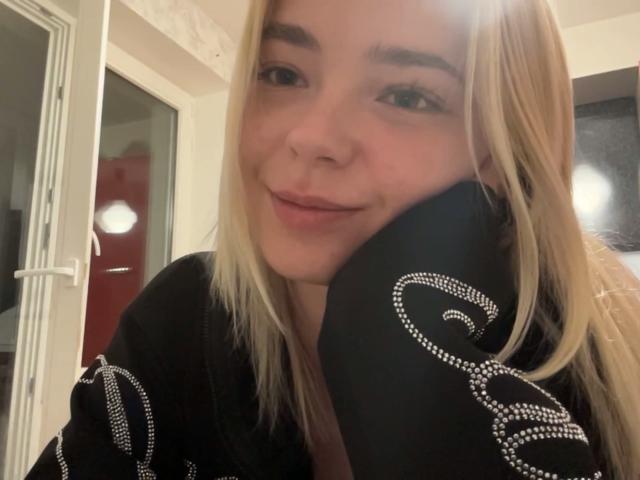 Adriene Degennaro, com cabelo loiro, um leve sorriso, camisa preta com padrões brancos em espiral, com o queixo apoiado na mão, em um selfie. Seu rosto está levemente avermelhado..