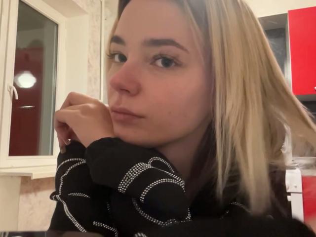 Adriene Degennaro mit blonden Haaren, einem schwarzen, gemusterten Hemd und einem nachdenklichen Gesichtsausdruck, die ihr Kinn in die Hand legt und zur Seite schaut, in einer schwach beleuchteten Küche. Ihre Nase ist klein..