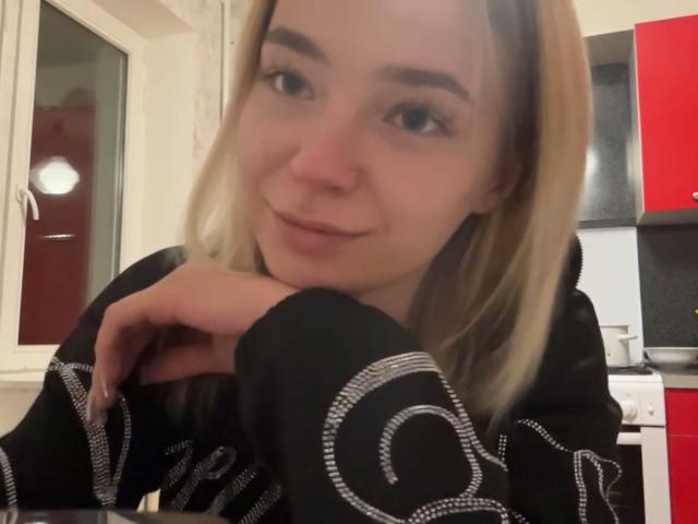 Adriene Degennaro mit blonden Haaren, einem schwarzen Pullover mit weißen Mustern, einem verschmitzten Lächeln, dem Kinn auf den Händen ruhend, blauen Augen und heller Haut, in einer Küche mit roten und schwarzen Schränken..