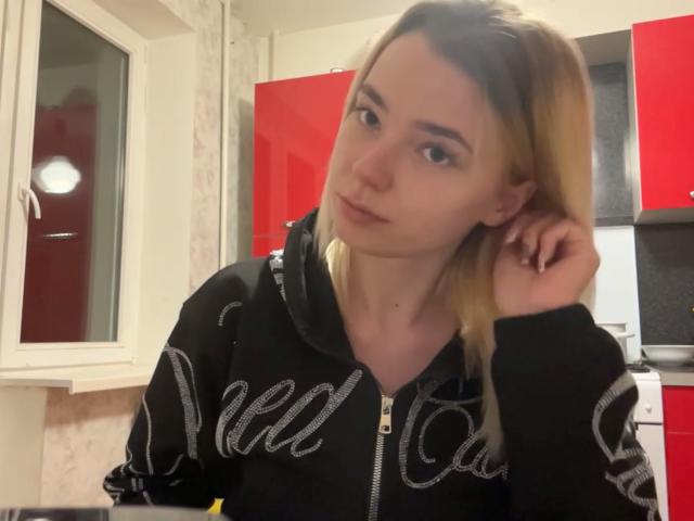 Adriene Degennaro, con cabello rubio, una sudadera negra de Nike y labios ligeramente fruncidos, apoya su barbilla en su mano en una cocina con armarios rojos. Parece aburrida..