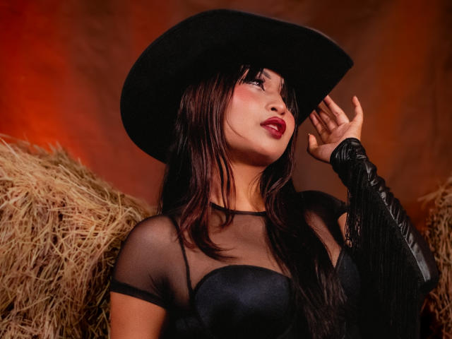 IssaJones with black hat, sheer top, and leather arm, looking up, has red lipstick, dark brown hair, and visible tits, standing near hay. Warm, orange-tinted background.