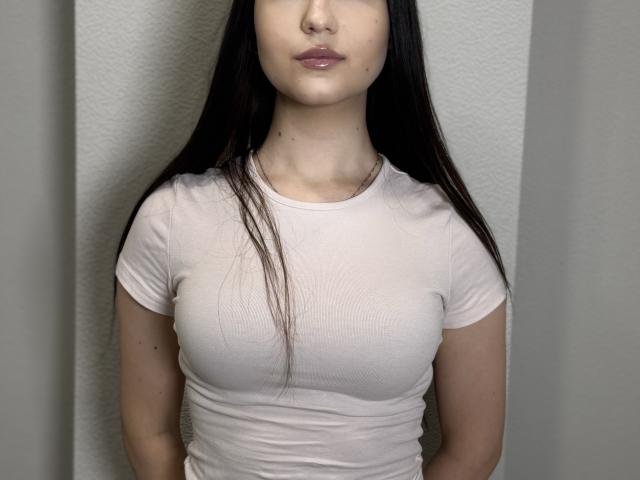 Anika Trentz, con cabello largo y oscuro, piel pálida, vestida con una camisa blanca ajustada, mostrando pechos pequeños pero notables, de pie frente a una pared lisa, con los labios ligeramente entreabiertos..