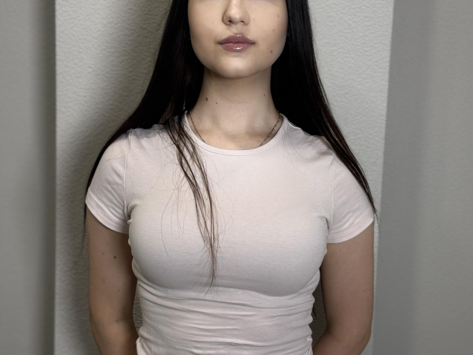 Anika Trentz, con cabello largo y oscuro, piel pálida, vestida con una camisa blanca ajustada, mostrando pechos pequeños pero notables, de pie frente a una pared lisa, con los labios ligeramente entreabiertos..