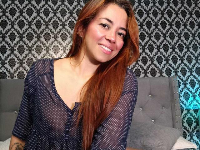 MadisonKoper with long red hair, smiling, wearing a sheer black top that shows her tits, standing in front of a gray tufted headboard.