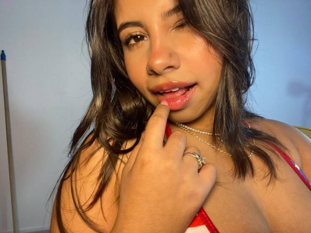 CelesteMorel with wavy brown hair, winking, touching her glossy pink lips with her finger, wearing a red top, gold necklace, and ring, showing her small, cute tits.