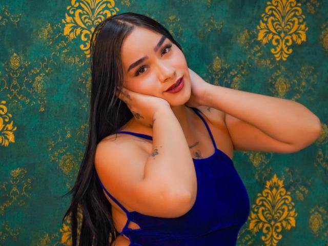 Vannesa Houston mit langem, schwarzem Haar, einem blauen Kleid und Tattoos, die ihr Gesicht mit den Händen berührt, lächelnd mit dunkelrotem Lippenstift, vor einem Hintergrund mit grün-goldenen Mustern..