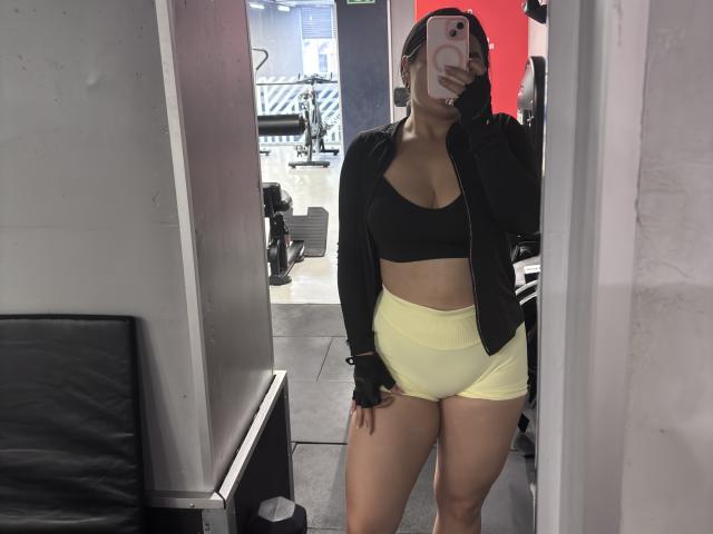 MiaDuboisX, die ein schwarzes Crop-Top, gelbe Hotpants und eine schwarze Jacke mit Reißverschluss trägt, macht ein Selfie vor einem Spiegel im Fitnessstudio und zeigt dabei ihre Brüste und ihren Hintern..