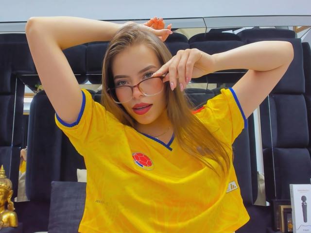 AnnaFer met een bril, een geel voetbalsok, blauwe accenten, een rood logo, handen omhoog, lang bruin haar, rode lippenstift, kijkt naar de camera, glimlachend, paarse nagels, gemiddelde borsten, binnen..