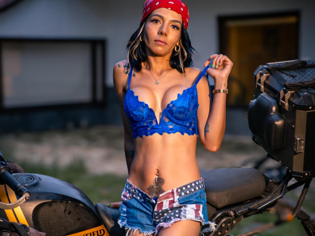 EvelinLuna, peau bronzée, soutien-gorge en dentelle bleu, petites seins, tatouage sur la hanche droite, short en jean avec drapeau américain, bandana rouge, et un vélo devant elle, tenant les bretelles du soutien-gorge..