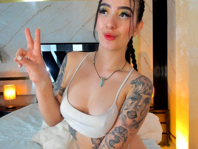 TifanyDiaz con tatuaggi, reggiseno bianco, che fa un gesto di pace, ombretto biondo, rossetto rosso, capelli neri intrecciati, collana con pendente verde, sorridente, letto sullo sfondo, illuminazione gialla tenue..