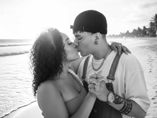 SolandAndy mit lockigem, dunklem Haar, küsst SolandAndy mit einer Mütze, die verkehrt herum getragen wird, beide halten Händchen, tragen Hosenträger und eine Uhr, beide haben Ohrringe, am Strand im Sonnenlicht..