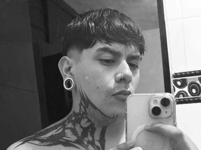 SolandAndy, con i capelli corti e scuri, un anello al naso e delle grandi espansioni alle orecchie, tiene in mano un telefono, mostrando un collo tatuato e guardando verso la fotocamera, in un selfie in bianco e nero..