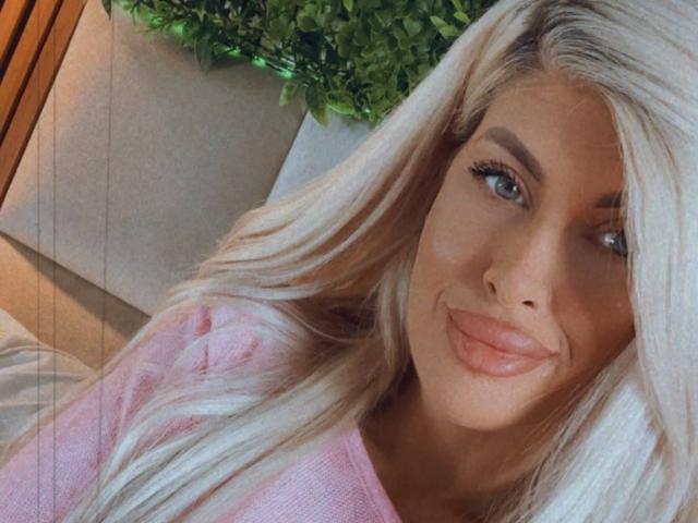 SalmaRoze mit langen blonden Haaren, vollen, rosafarbenen Lippen und blauen Augen, trägt ein rosa Oberteil, lächelt leicht und schaut in die Kamera. Ihre Brüste sind nicht sichtbar..