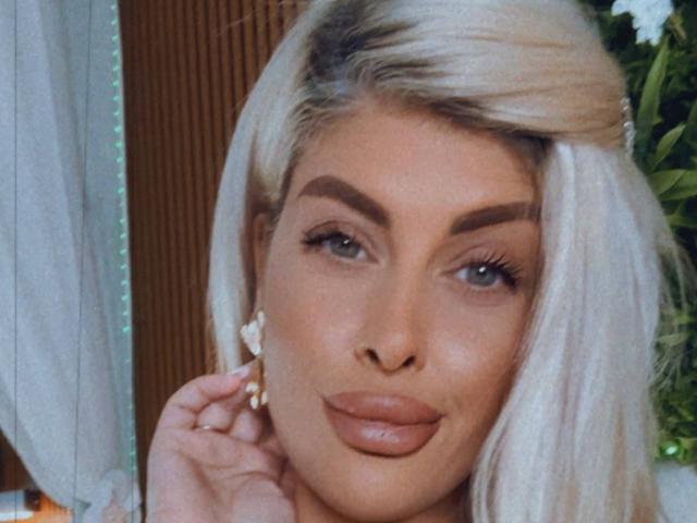 SalmaRoze has platinum blonde hair, full eyebrows, blue eyes, and glossy brown lips. She's touching her ear with a hand, wearing a floral earring.
