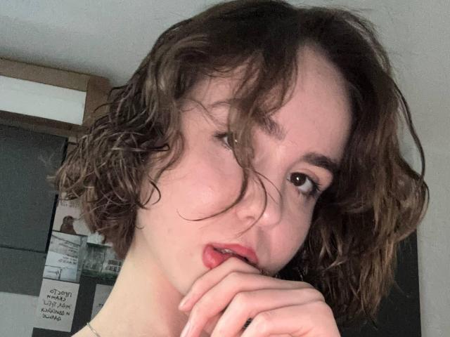 NylaDoore, con i capelli castano disordinati, la pelle pallida e le labbra rosse, morde il dito con la bocca leggermente aperta, guardando nella fotocamera. Ha piccoli seni e occhi marroni..