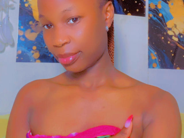 JazzBerry tiene la piel morena, pechos pequeños y un top rosa sin tirantes. Está sacando la lengua con una expresión juguetona, lleva el pelo trenzado y un pendiente de diamantes..