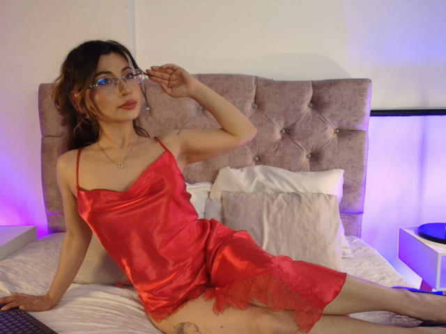 SeleneLeon con biancheria intima di seta rossa, occhiali e rossetto rosso, seduta su un letto, che mostra il sedere e un piccolo tatuaggio sulla coscia. Camera da letto con luci rosa e viola..