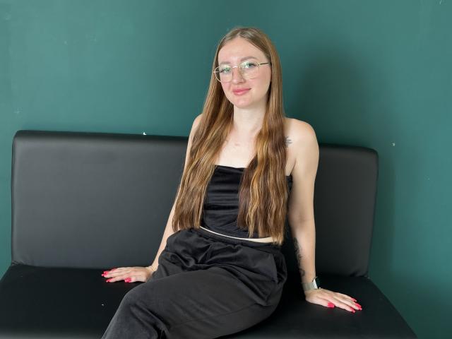 AnnaMillsk, com cabelos castanhos longos, óculos, blusa preta sem alças, calças pretas, sentada em um sofá preto, sorrindo, unhas vermelhas, pequena tatuagem no braço direito, parede de fundo verde..