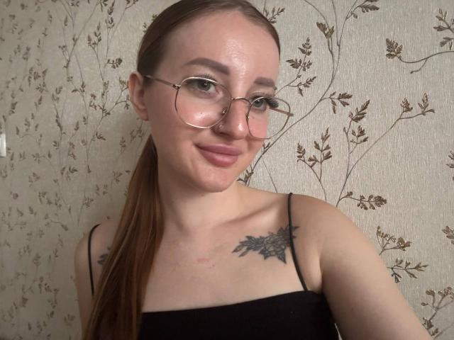 AnnaMillsk mit Brille, lächelnd, trägt ein schwarzes Top mit Spaghetti-Trägern, zeigt ein Brust-Tattoo, hat braunes Haar im Pferdeschwanz, helle Haut und eine Nasenpiercing..