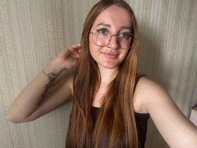 AnnaMillsk mit langen braunen Haaren, Brille und Nasenring, lächelnd in die Kamera, trägt ein schwarzes Tanktop und zeigt ein Tattoo auf ihrem rechten Arm..