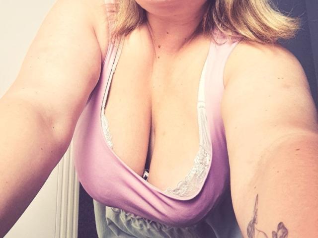 RadaRori, avec de grandes seins pendants, porte un débardeur rose laissant apparaître une bordure en dentelle blanche, a les cheveux blonds et un tatouage sur son bras droit, et elle prend un selfie..