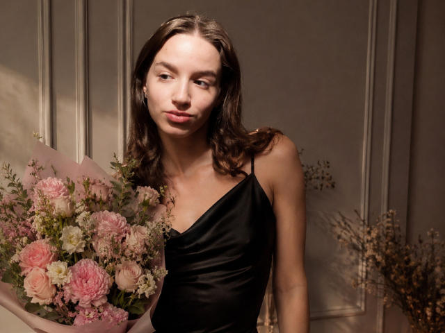 Kleral, avec ses cheveux bruns ondulés, portant une robe en satin noir, tenant un bouquet de fleurs roses et blanches, affichant un léger sourire, dans une pièce éclairée doucement..